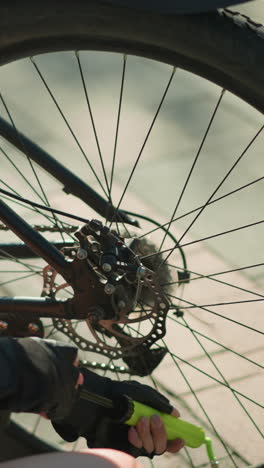 close-up back view of person squatting to pump air into bicycle tire using green nozzle, with black gloves on hand and blurred background of wheel spokes, pavement, and soft shadows