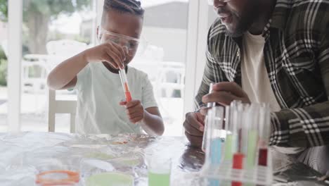African american father and son sitting at table holding test tubes with liquid, in slow motion