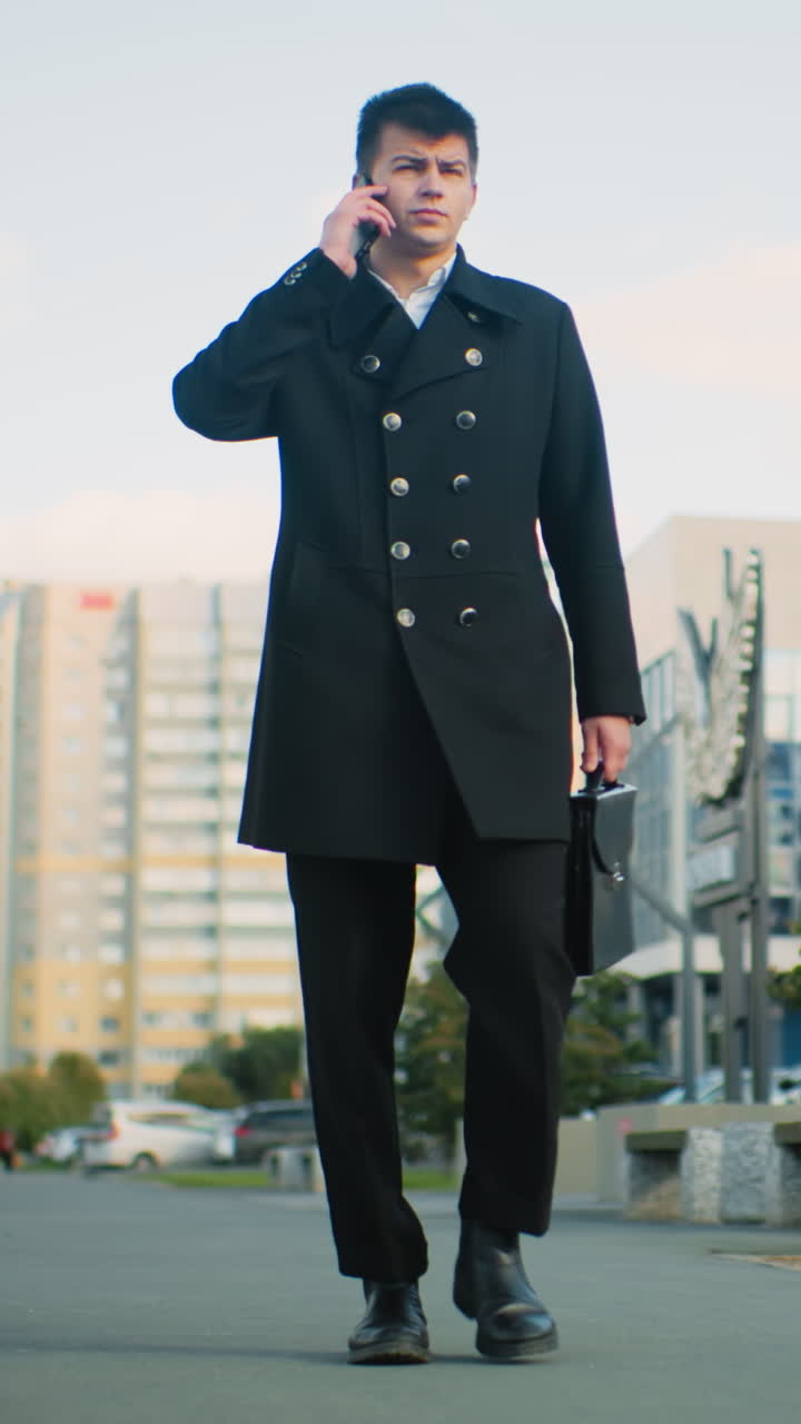 검은 코트를 입은 선생님은 밖에서 자신감 있게 고, 은 낮빛 아래 도시 초록색과 흐릿한 현대 건물로 둘러싸인 전화를 받고 전화에 응답하기 위해 주머니에 손을 고 있습니다.