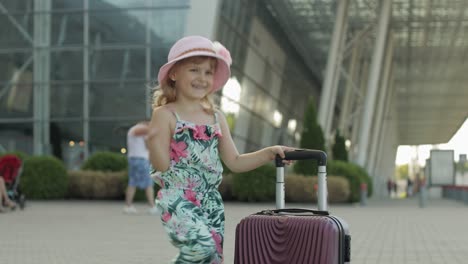 child girl tourist with suitcase bag near airport. kid with luggage dances, rejoices, show thumbs up