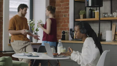 chica con auriculares desayunando y trabajando en un ordenador portátil en un piso compartido