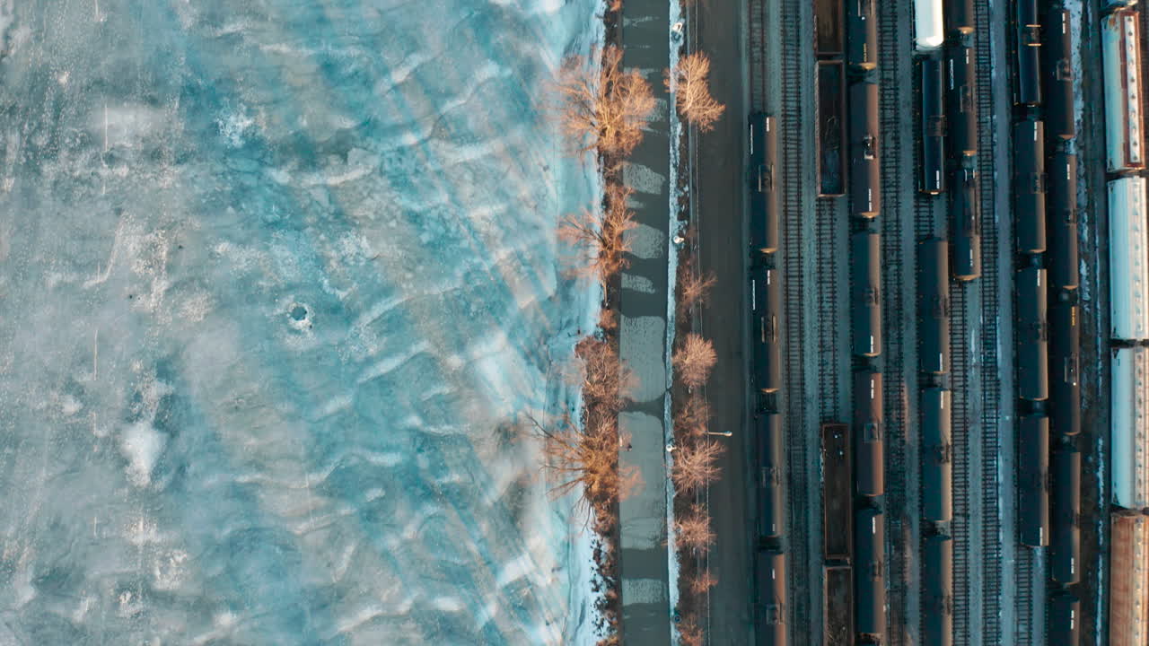 Ripresa aerea di scali ferroviari e sentiero pedonale lungo un fiume ghiacciato.