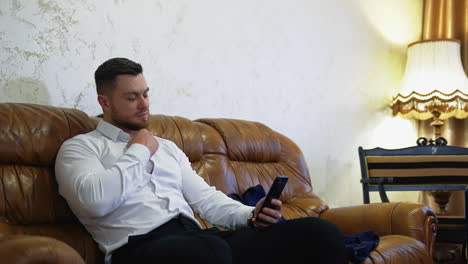 handsome man on couch with a mobile phone. young businessman sitting on a sofa in a hotel and unbuttoning his white shirt while viewing in the phone.