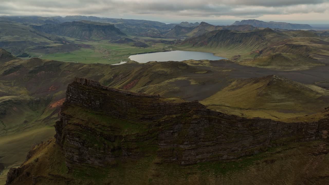 Aerial view of a serene lake surrounded by volcanic highlands and rugged cliffs near Hafursey, Iceland, under overcast skies.