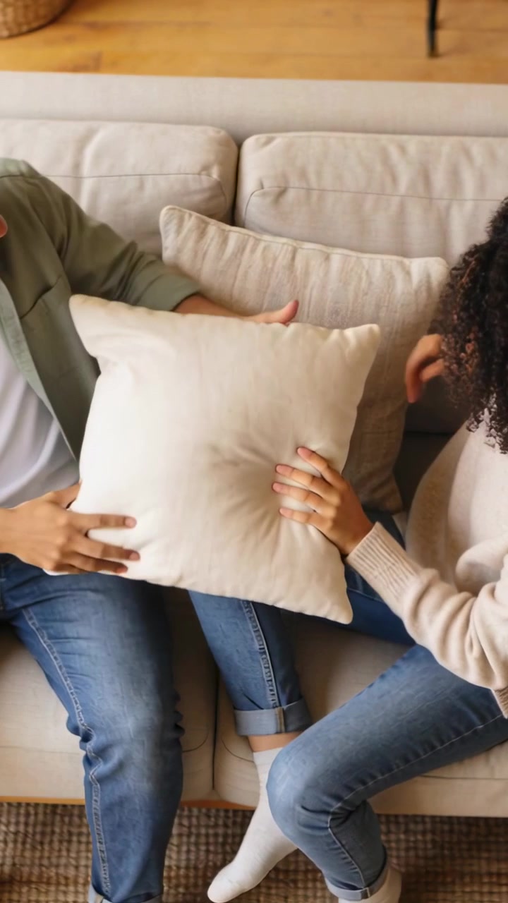 Video verticale: Donna sorridente che allunga la mano verso un cuscino, uomo che la imita e tira giocosamente sul divano di casa.