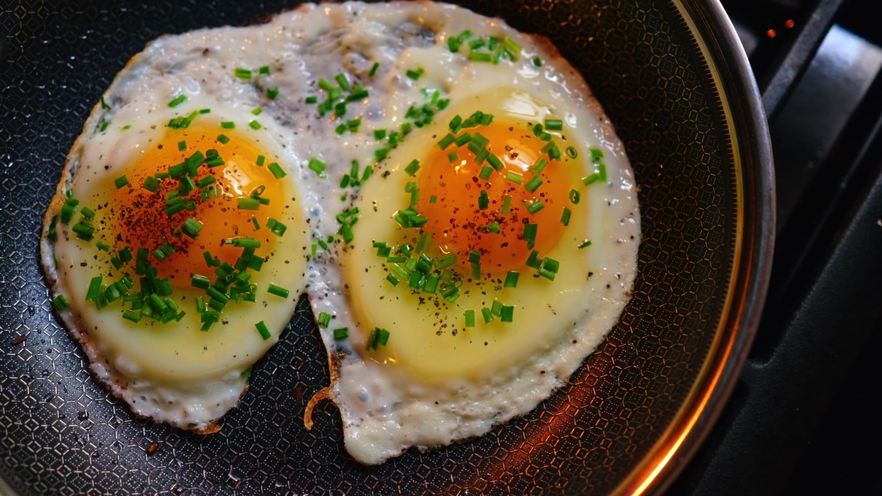 真上から見た、チャイブが添えられた目玉焼き2つ。刻んだチャイブと胡椒が振られた目玉焼き2つが、質感のある六角形の焦げ付きにくいフライパンに入っているハイアングルクローズアップ。