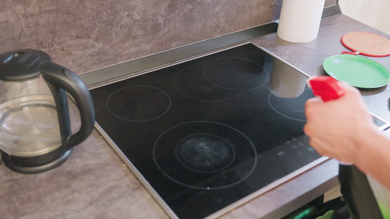 Close up hand holding red spray bottle applies cleaning liquid on black glass cooktop before wiping surface with green cloth in modern kitchen, kettle and plate visible on countertop