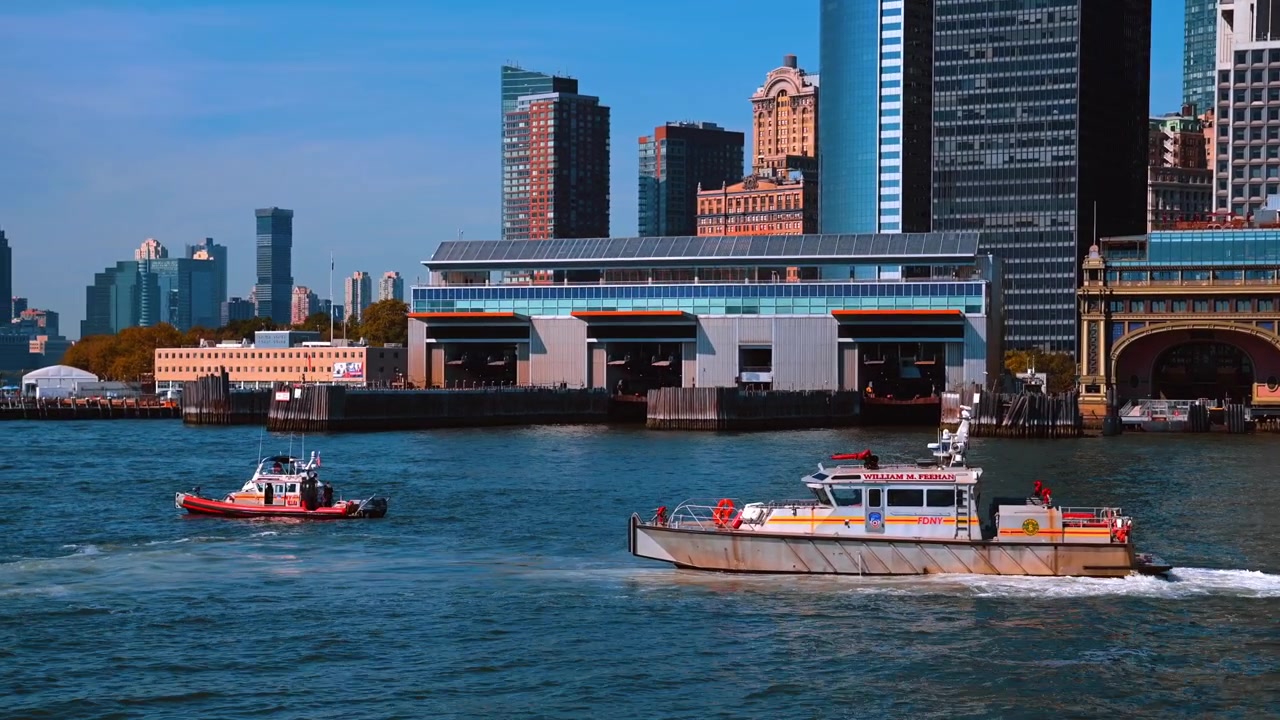 New York, USA, 4 May 2026 : FDNY fireboat passes Battery Maritime Building in Lower Manhattan. Silver rescue vessel creates a wake beside another boat, with arched ferry slips and towers in sunlight.