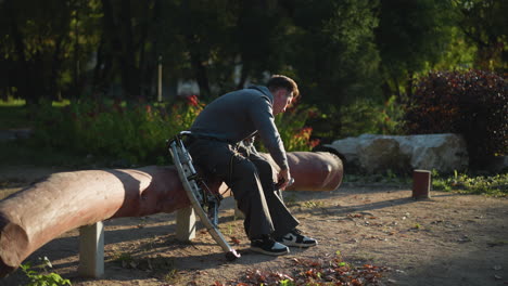 chap sentado en madera al aire libre en sudadera de capucha gris oscuro y pantalones de sudadera, intensamente enfocado en el ajuste de pilotes cargados de resorte. rodeado de árboles y vegetación en un entorno natural pacífico