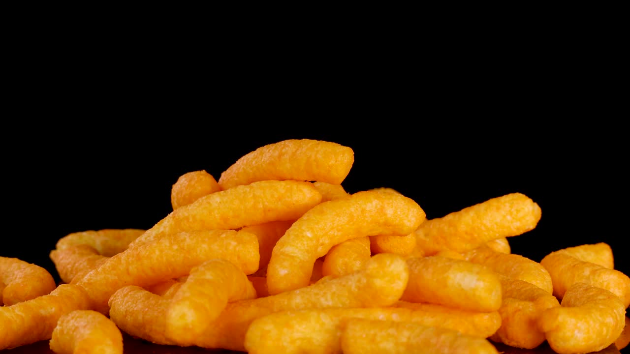 Crunchy cheese puffs fall onto a pile, brightly lit against a dramatic black background, close-up