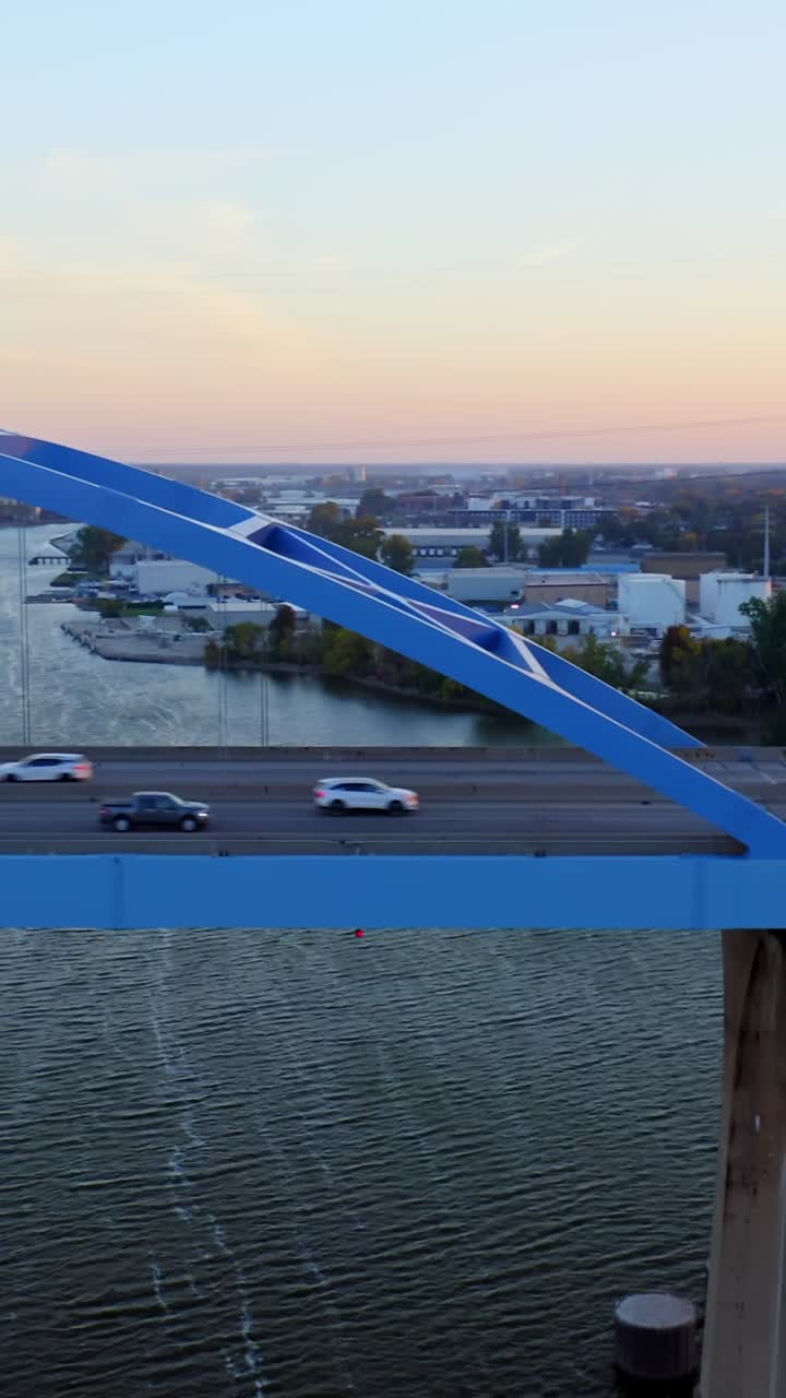 A red car moves across a bright blue steel bridge above the river as the golden sun sets behind distant tanks and trees, casting warm reflections on the rippling water below