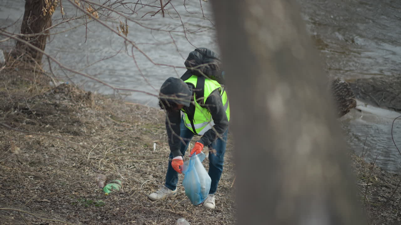 Volunteer in reflective vest, hoodie, and backpack wearing gloves collects plastic waste from ground near riverbank, placing litter into blue bag during environmental cleanup effort to protect nature