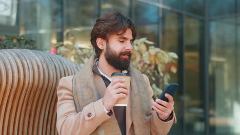 happy indian young man tourist enjoying morning coffee hot drink sitting on city downtown street