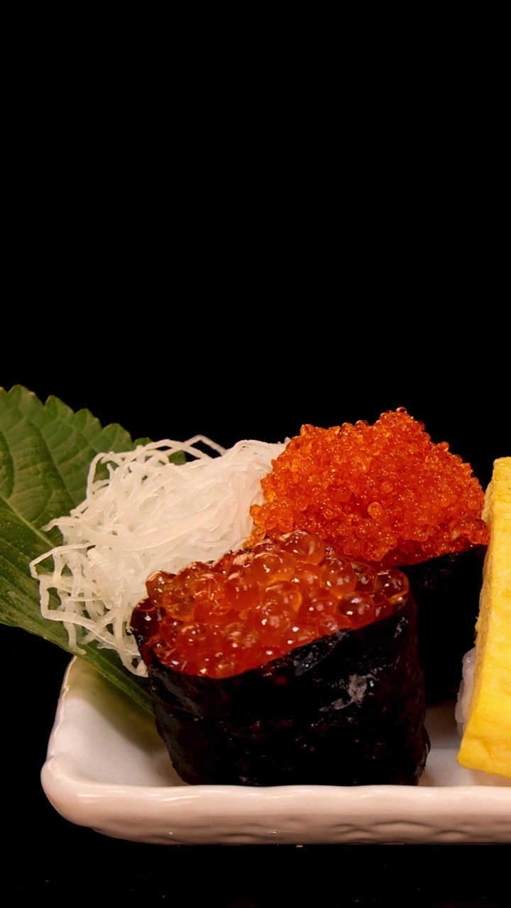 Un piatto rettangolare bianco con nigiri, sashimi e roll di sushi assortiti ruota lentamente sotto una brillante illuminazione da studio su uno sfondo nero, mettendo in risalto frutti di mare e guarnizioni vivaci