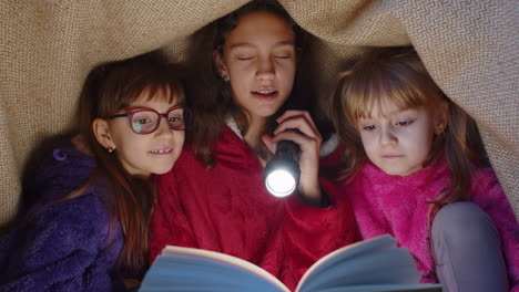 sisters girl kids under blanket reading interesting fairytale story book using flashlight at home