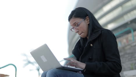 Mujer-Hindú-Sonriente-Con-Anteojos-Escribiendo-En-Una-Computadora-Portátil-Al-Aire-Libre