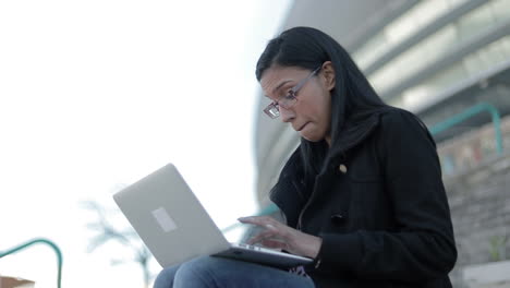 smiling hindu woman in eyeglasses typing on laptop outdoor