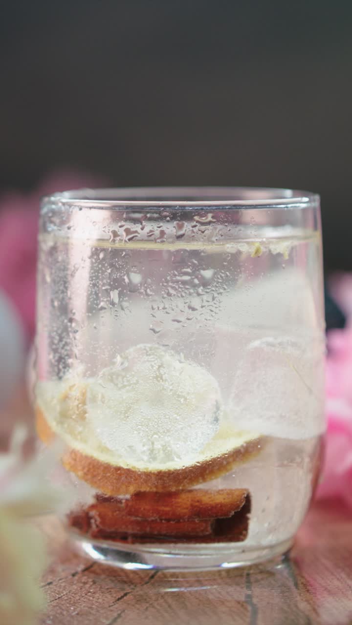 Pan shot of citrus drink with ice cubes, lemon slice, and cinnamon sticks in clear glass on wooden surface