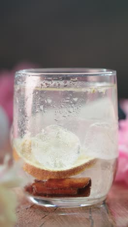 pan shot of citrus drink with ice cubes, lemon slice, and cinnamon sticks in clear glass on wooden surface