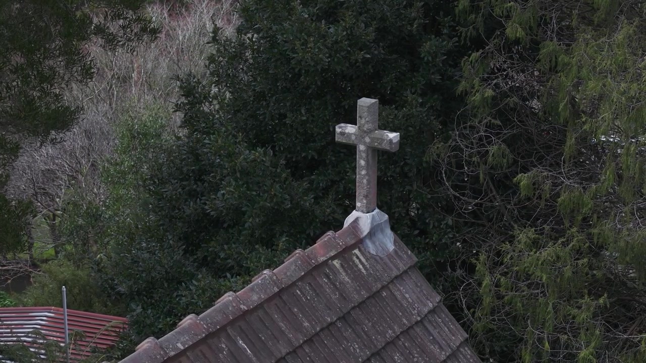 Scatto grandangolare del tetto in tegole consumato di una vecchia chiesa con una croce di pietra a Olinda, Melbourne, incorniciato da una fitta vegetazione e una luce naturale suggestiva, senza persone visibili.