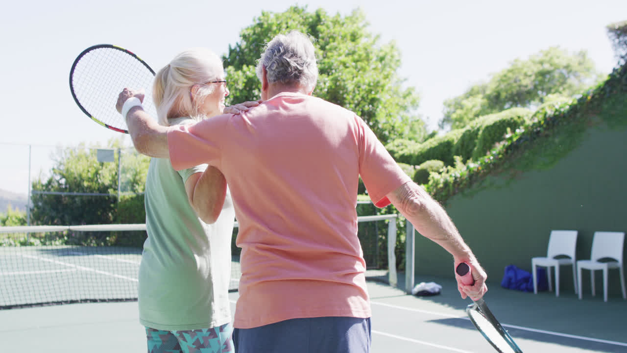Smiling senior caucasian couple with rackets, embracing at sunny outdoor tennis court, slow motion