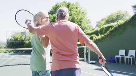 una pareja caucásica senior sonriente con raquetas, abrazándose en la soleada cancha de tenis al aire libre, en cámara lenta