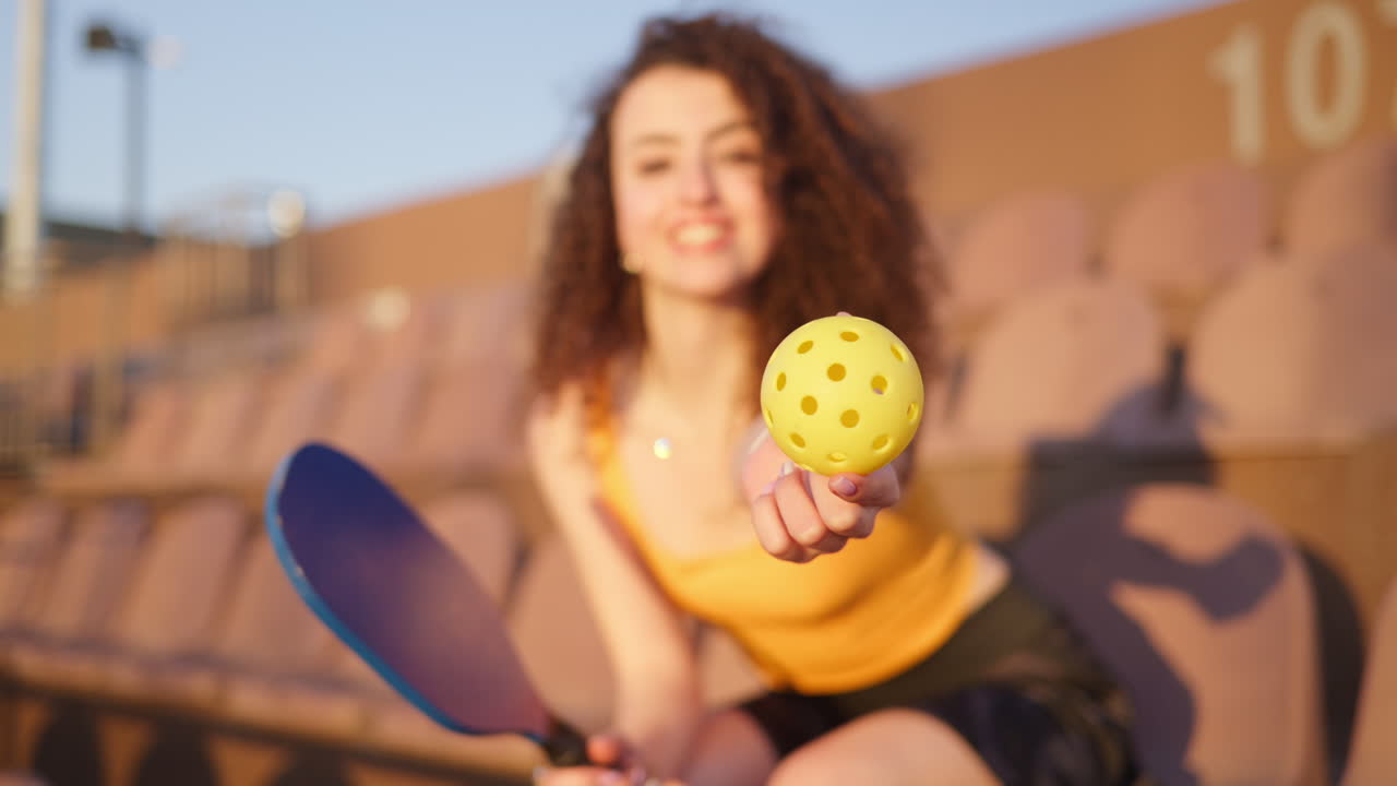Close up of a yellow ball with a blurry view of a woman with curly hair sitting on a courtside seat, holding a pickleball racket