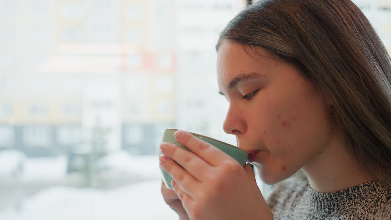 Young freelancer enjoying hot coffee by window during snowy day, gazing outside with calm expression. Warm indoor ambiance contrasts with cold winter view, highlighting peaceful, cozy work-from-cafe