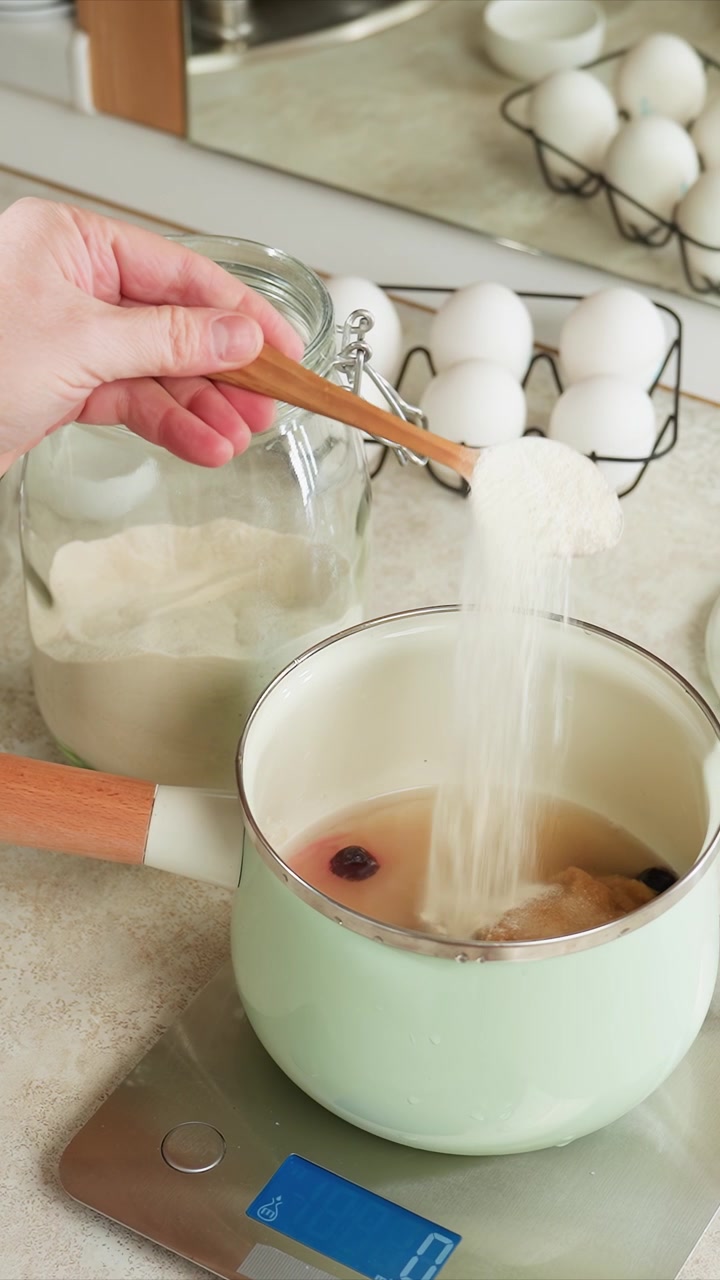 Hand scoops white powder from glass jar with wooden spoon. Glass jar contains light colored powder and wooden spoon. Kitchen countertop with glass jar and digital scale