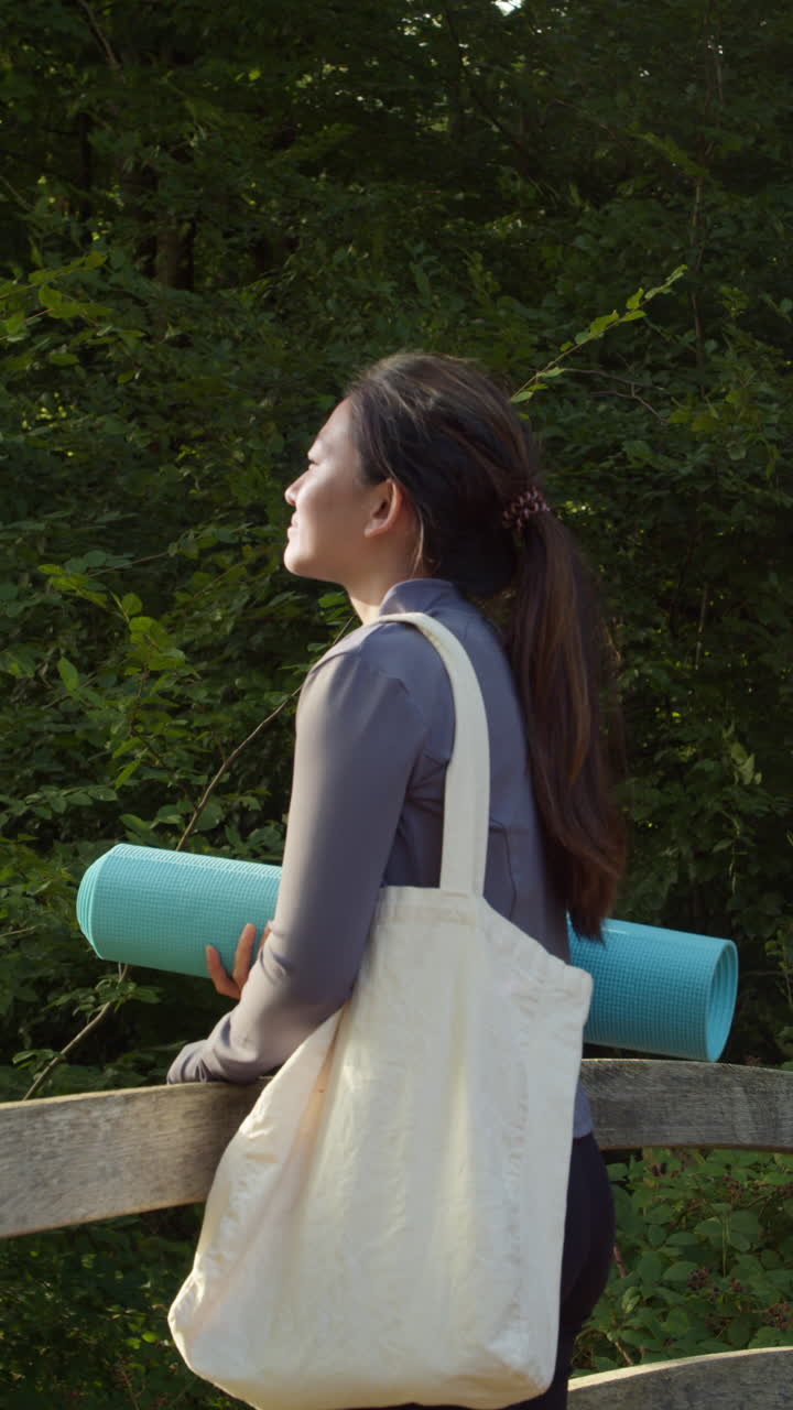 video verticale di una donna che indossa abiti sportivi che porta un tappetino da yoga in piedi su un ponte nella foresta che si guarda intorno godendo la pace e la bellezza della natura girato in tempo reale