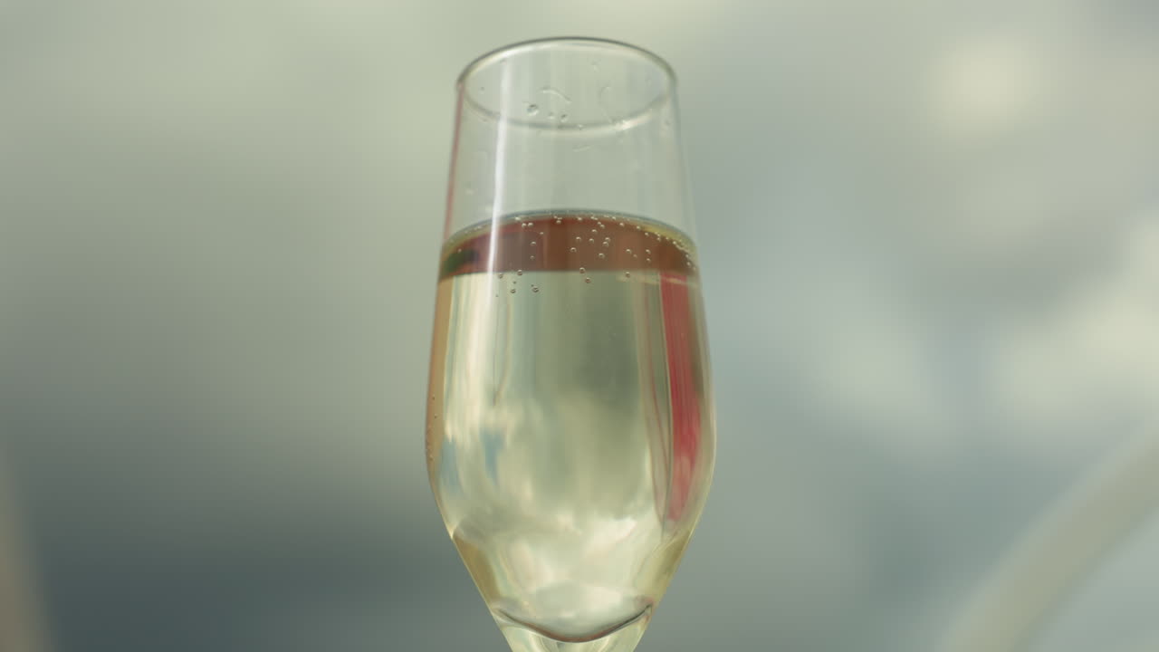 Calice da champagne colorato in stile carnevalesco con riflesso di tessuto rosso, primo piano di un flûte di prosecco che mostra le bollicine che salgono, cielo nuvoloso luminoso sullo sfondo, atmosfera festosa e allegra con calda luce solare e ridotta profondità di campo
