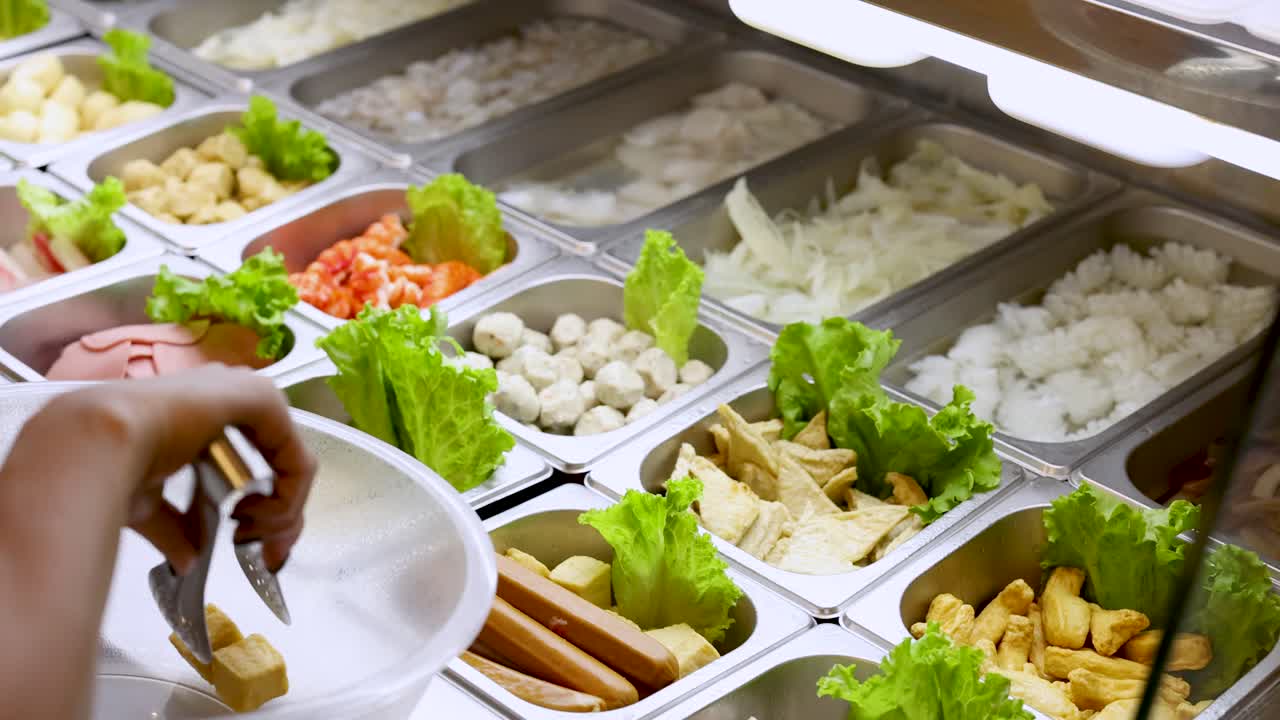 Person uses tongs to pick tofu and vegetables from well-lit Asian hotpot buffet counter