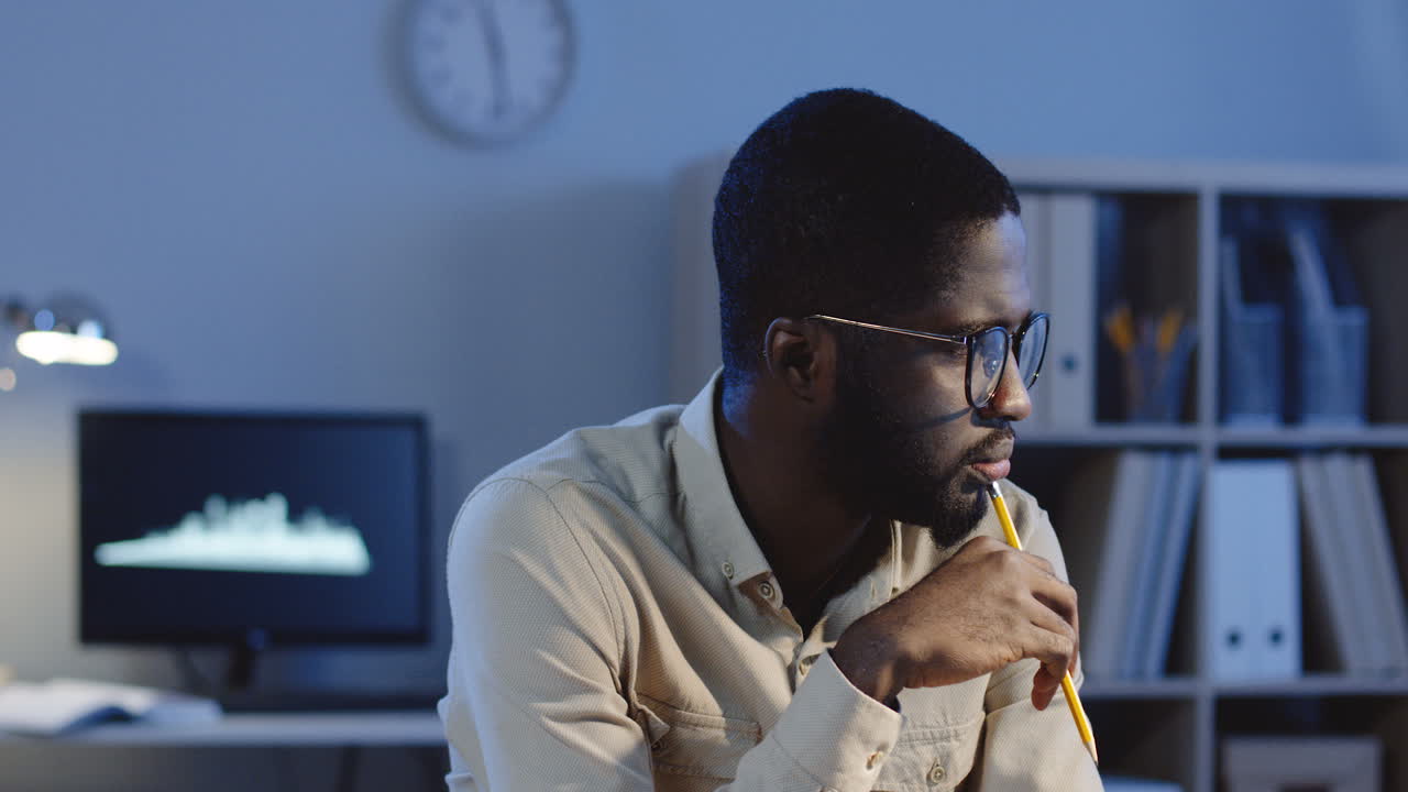 Portrait Of Serious Man In Glasses Looking At The Side, Thinking And Spinning A Pencil In A Hand In The Office At Night