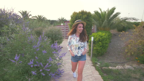 a young woman in a floral shirt and denim shorts smiles while walking down a path in a garden on a sunny day