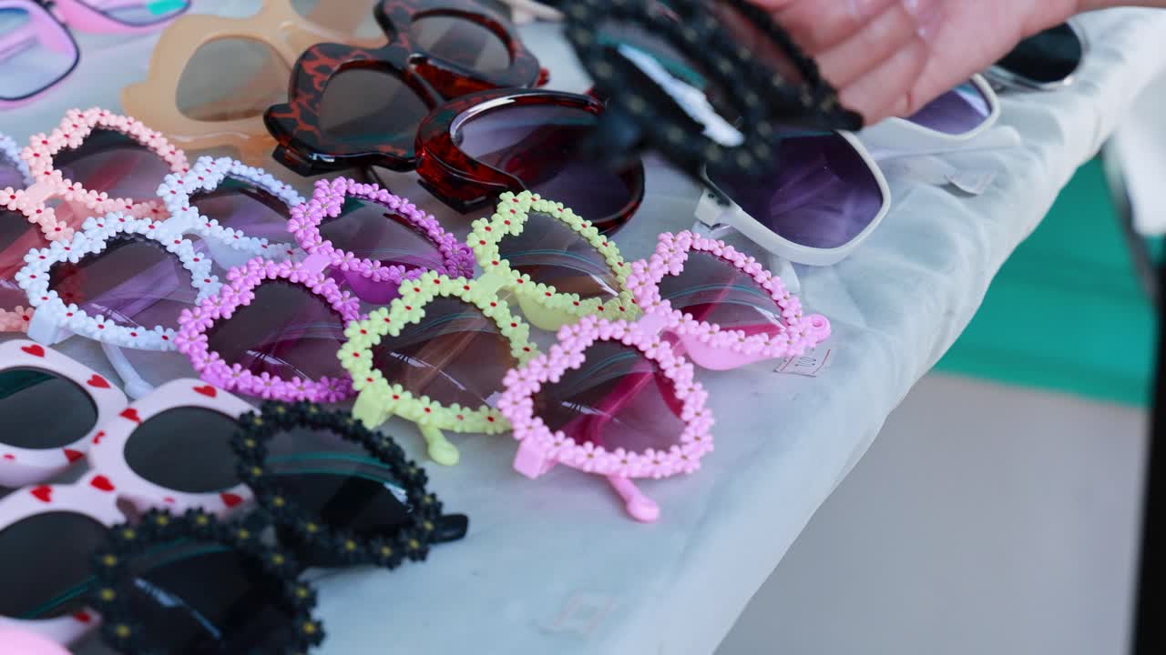 A person examines colorful sunglasses on a market stall in Phuket, Thailand, under bright lighting with close-up camera angles