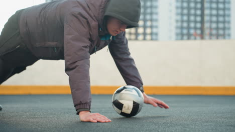 deportista realizando flexiones alternando la colocación de la mano en la pelota de fútbol mostrando fuerza, coordinación y agilidad en una arena deportiva urbana al aire libre con un fondo de edificio residencial moderno