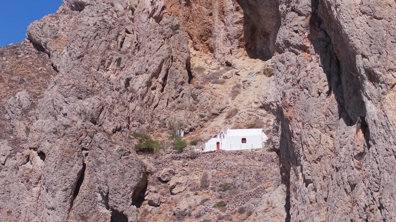 Ripresa con drone che rivela una cappella situata tra le rocce sopra la spiaggia di Perissa a Santorini, mostrando drammatiche scogliere vulcaniche e lo splendido paesaggio costiero dell'Egeo