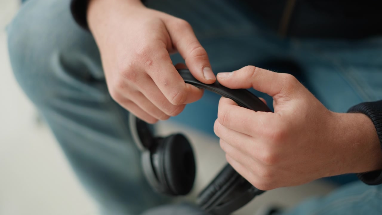 Tampilan dekat tangan menyesuaikan headphone over-ear hitam yang berfokus pada ikat kepala dan bantalan telinga. Orang tersebut mengenakan celana jins biru dengan atasan gelap yang terlihat dalam pencahayaan dalam ruangan yang lembut. Latar belakang buram.