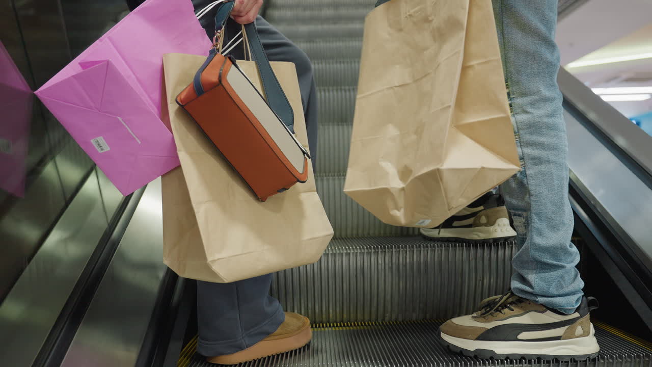 Leg view of two people ascending escalator while carrying paper shopping bags, gift bags, and handbag in modern shopping mall with reflective surfaces and bright lighting