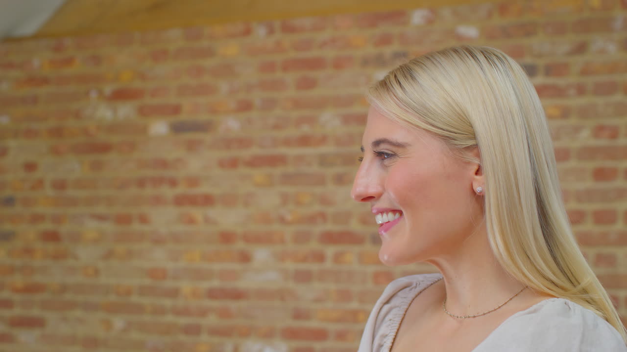 joven de pie junto a una pared de ladrillo en casa hablando y riendo con una persona fuera de la cámara