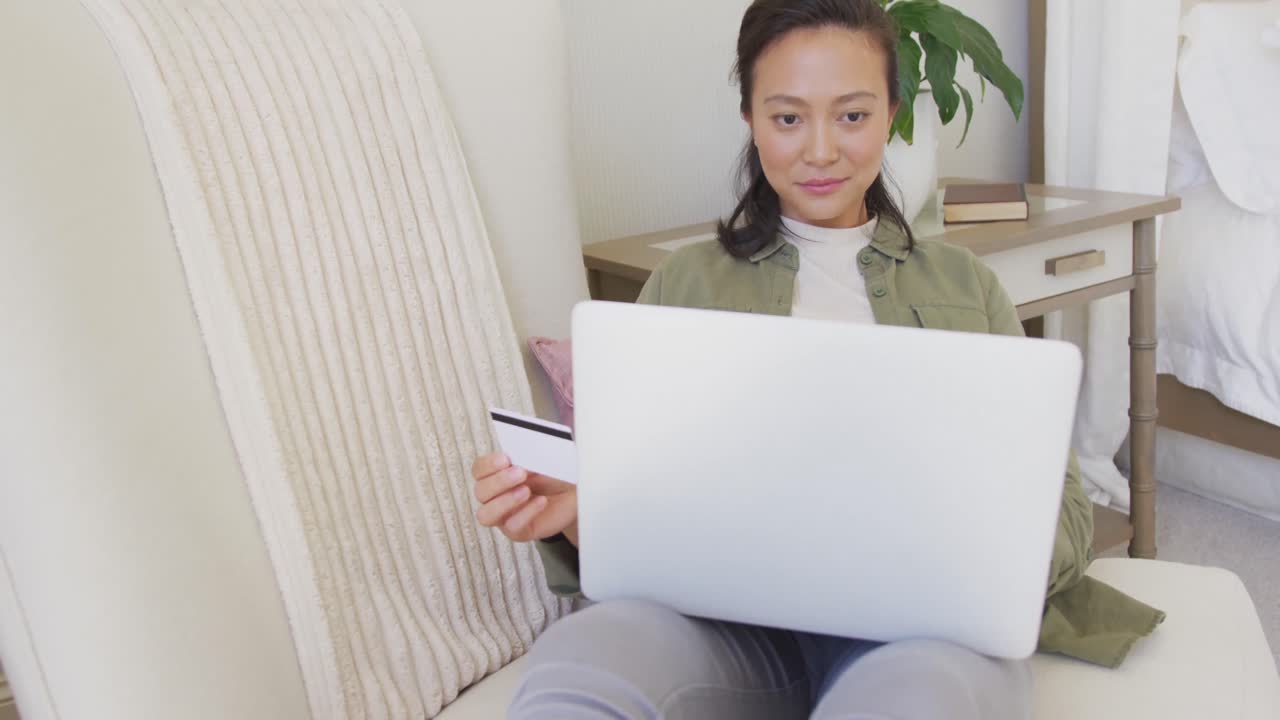 快乐的亚洲女性在卧室里使用笔记本电脑并拿着信用卡，慢动作。
