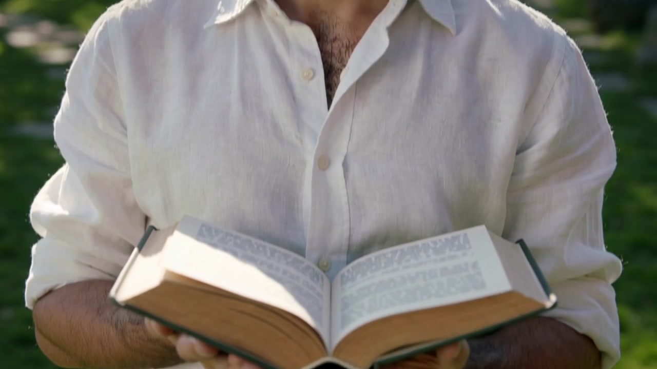 La telecamera inquadra un uomo in camicia di lino che tiene in mano un libro rilegato aperto su un sentiero in giardino, presentandolo alla telecamera.