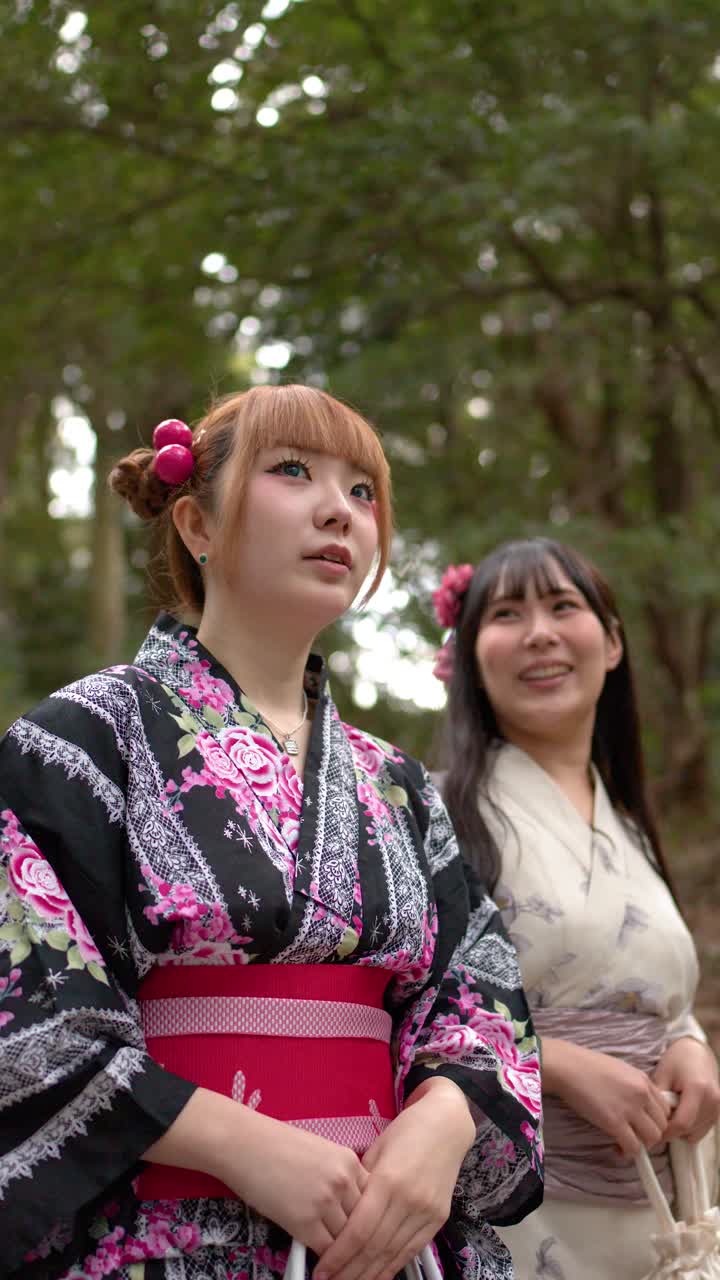 Dua teman mengenakan kimono bermotif bunga berjalan melalui taman hutan yang rimbun di bawah cahaya alami yang lembut.