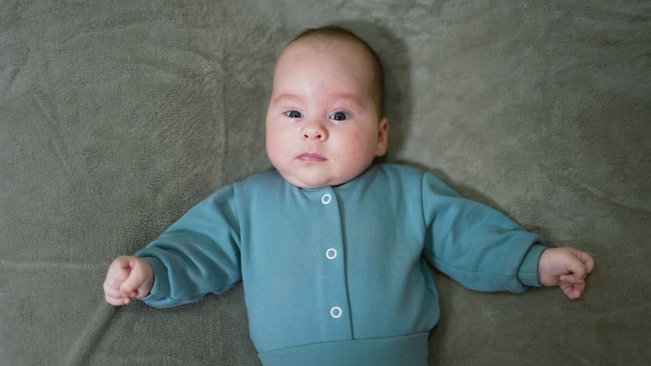 Baby boy wearing blue warm suit lying on the soft bed. Child moving his hands actively. Grey blanket background.