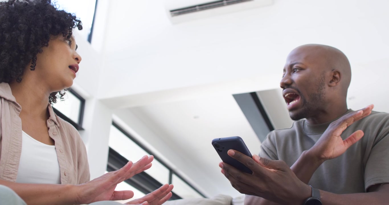 Afroamerikansk par på sofa derhjemme, mand viser smartphone, kvinde reagerer og diskuterer.