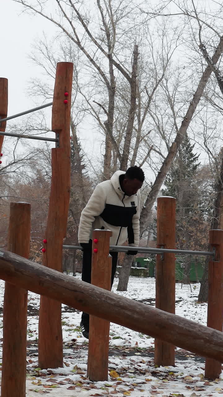 hombre negro sube en equipos de entrenamiento al aire libre durante el invierno, realizando ejercicios de empuje mientras cae, rodeado de tierra cubierta de nieve y árboles desnudos en el fondo