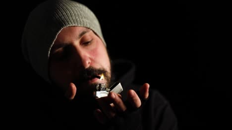 close up of a contrasted portrait of young man with beard and hat lighting cigarette and looking to camera