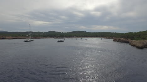 Drone-shot-flying-over-a-bay-in-Ibiza-Spain-with-boats-floating-around-on-a-cloudy-day-near-the-shore-and-sea-LOG