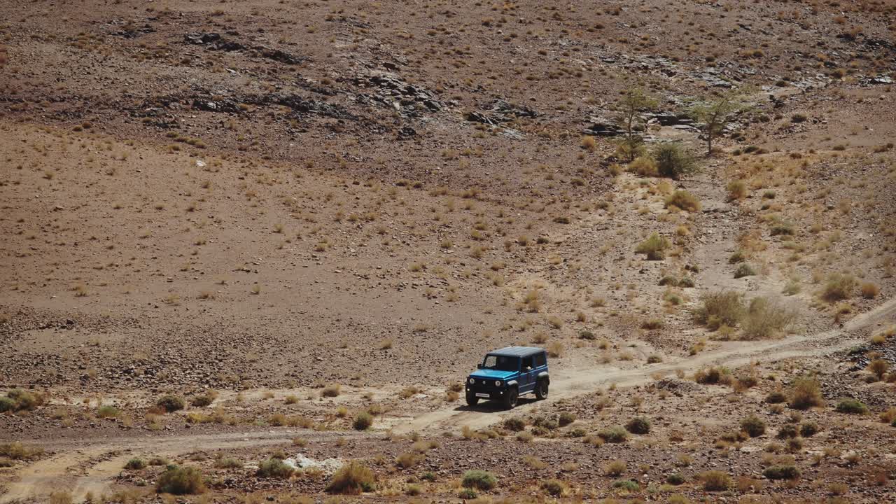 Veicolo fuoristrada che guida attraverso l'arido terreno desertico marocchino sotto un cielo limpido, panoramica media con teleobiettivo che oscilla sulla strada