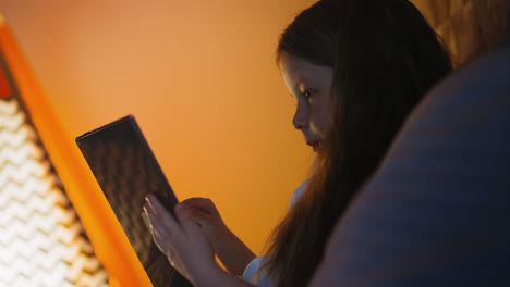 una chica navega por internet en una tableta apoyándose en almohadas en la cama.
